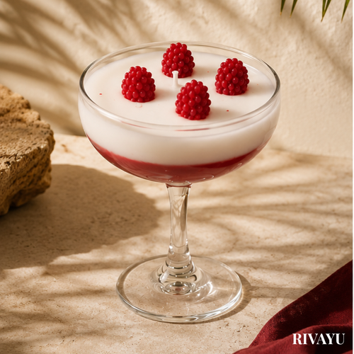 A dessert-style glass candle in a coupe-shaped stemware bowl, filled with white wax and a red berry-colored layer at the bottom, topped with four red raspberries. The glass is photographed on a beige stone surface with warm sunlight and shadows, and the brand name RIVAYU appears in the bottom right corner.