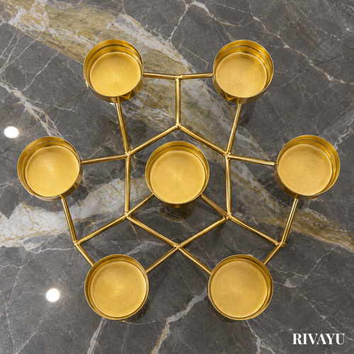 A gold-finished metal 6-candle holder with a geometric web-like frame, photographed from directly above on a dark gray marble surface. The brand name 'RIVAYU' is visible in the bottom right corner.